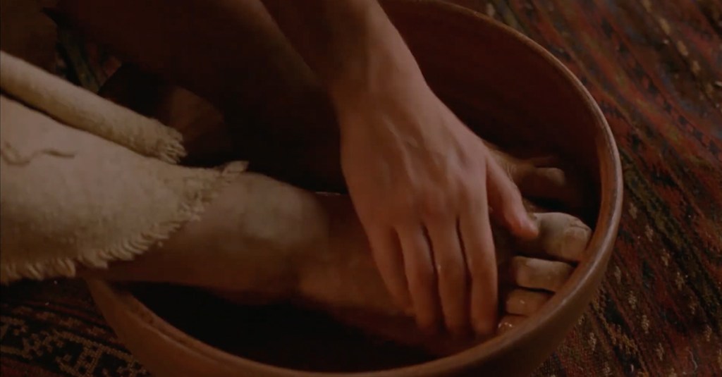 A close-up image showing a pair of hands gently washing someone else's dusty feet, which are placed inside a simple wooden bowl. The bowl rests on a colorful, patterned woven rug, evoking a sense of humility and service.