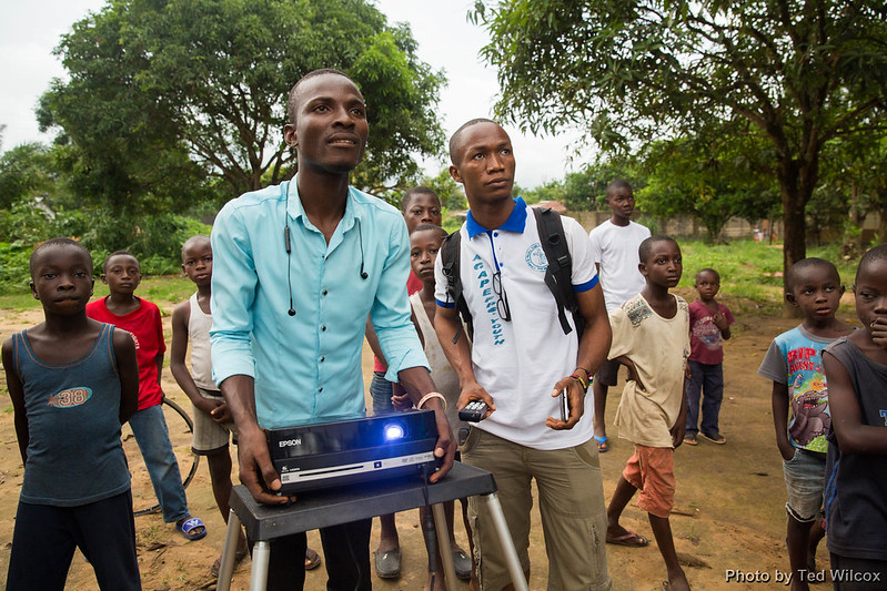 Man holding a projector and another man holding a remote surrounded by village children
