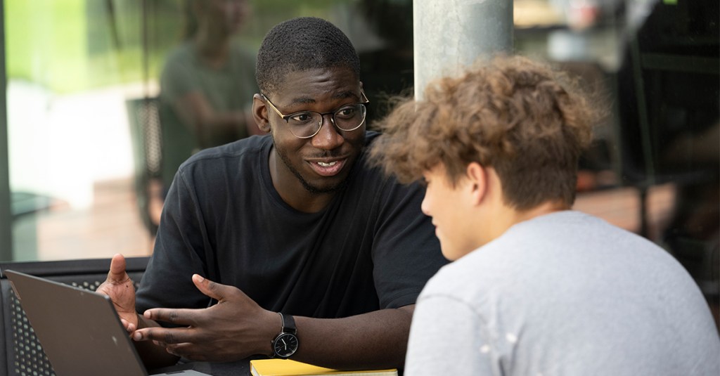 Two college-age students having an earnest conversation outdoors, potentially discussing apologetics and evangelism while looking at notes on a laptop.