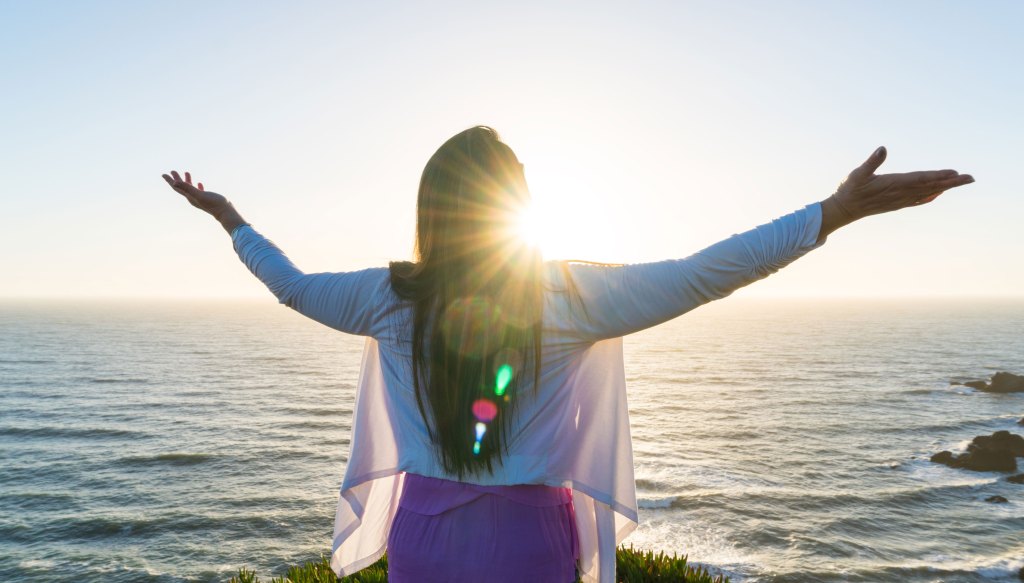 A woman standing at the edge of a coastline with her arms outstretched, facing the ocean as the sun sets behind her, creating a bright sunburst effect.