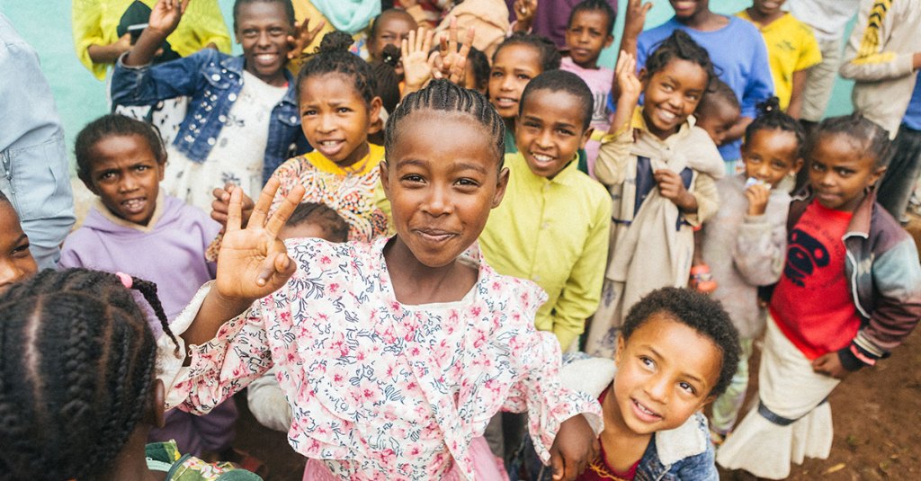 A large group of children are gathered closely together outdoors, smiling and looking at the camera in front of a bright teal wall. In the center foreground, a young girl in a floral dress smiles warmly and holds up two fingers in a peace sign. Many other children surround her, some smiling and waving.