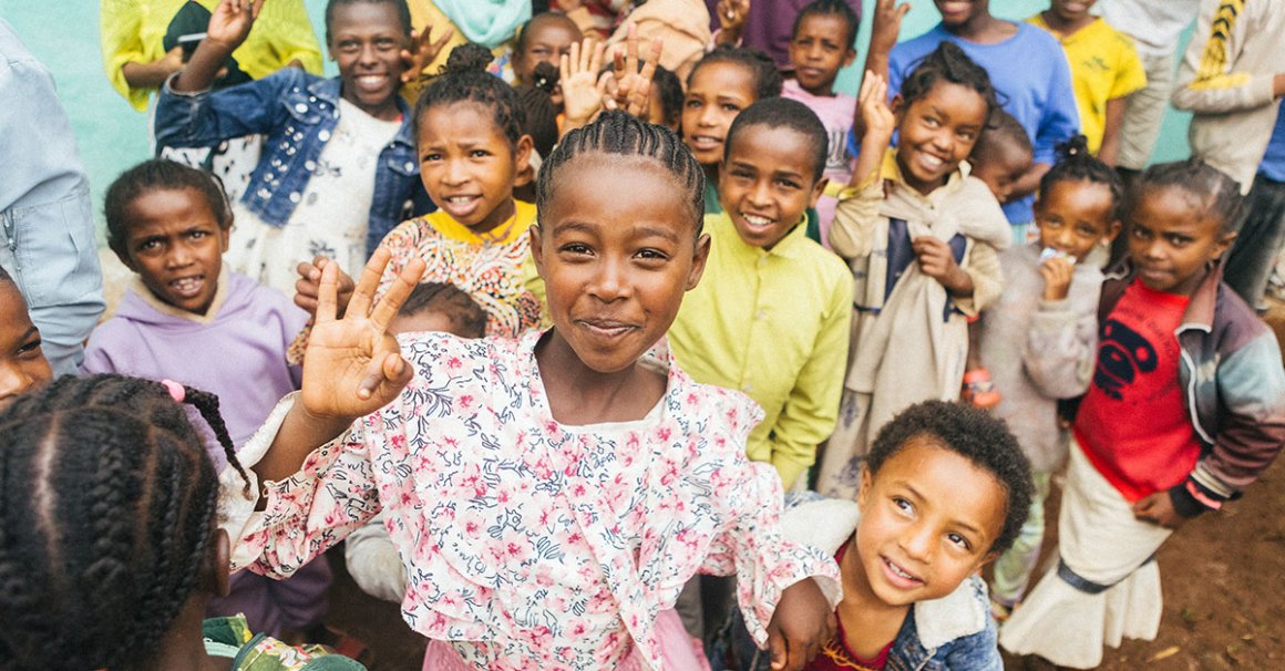 A large group of children are gathered closely together outdoors, smiling and looking at the camera in front of a bright teal wall. In the center foreground, a young girl in a floral dress smiles warmly and holds up two fingers in a peace sign. Many other children surround her, some smiling and waving.