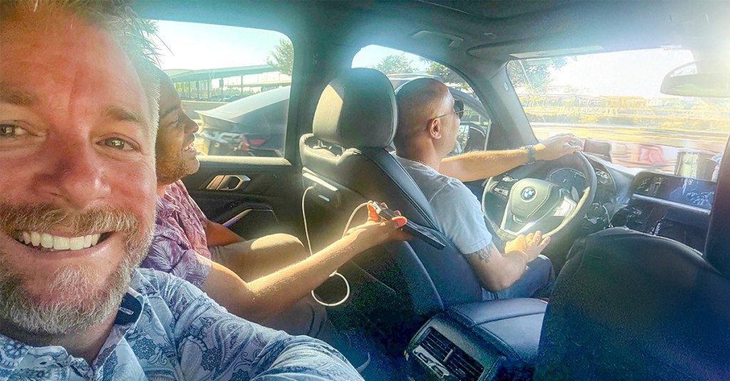 A smiling, bearded man takes a selfie from the front passenger seat of a car. A bald man wearing sunglasses is in the driver's seat, and another person is visible smiling in the backseat. Bright sunlight fills the car.