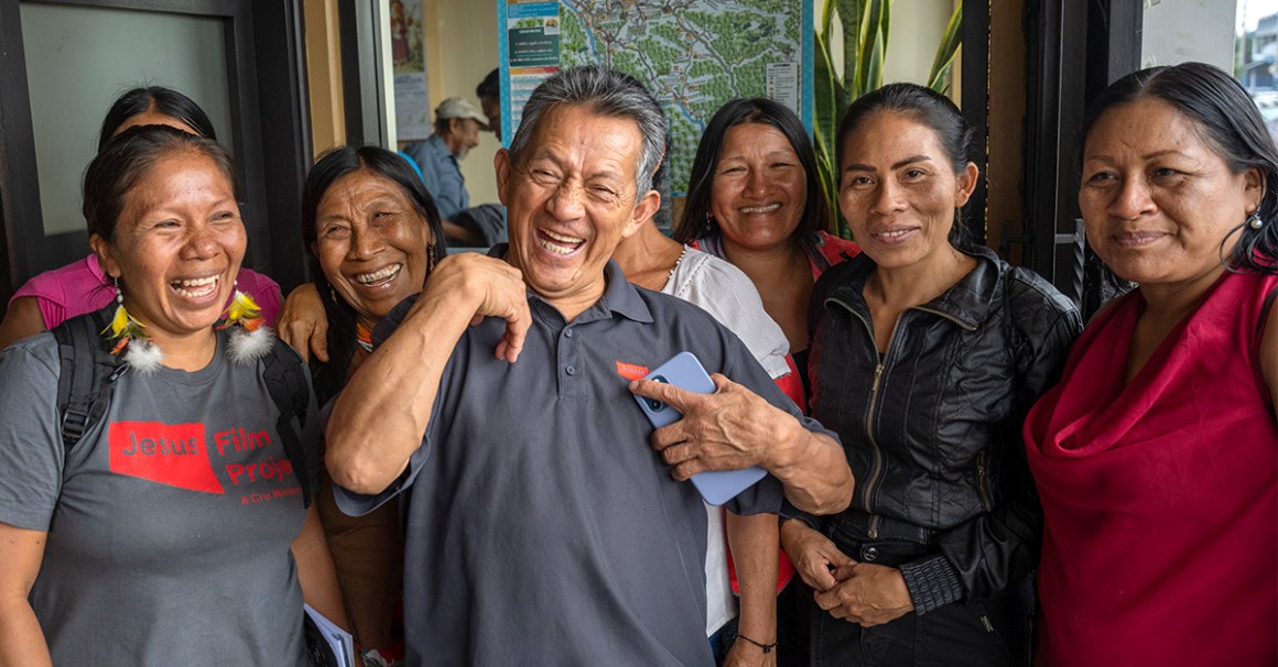 A group of six adults stand closely together indoors, smiling and laughing warmly. A man in the center holds a phone and grins broadly, while the others—mostly women—gather around him, some wearing casual clothing and one in a shirt that reads “Jesus Film Project.” A colorful map is visible on the wall behind them, and the mood is joyful and communal.