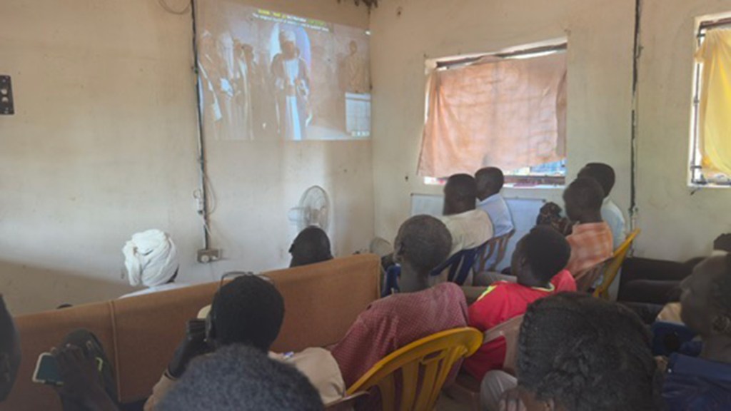 A group of people intently watches the JESUS film in a small room. The film is being projected on a white wall. This is an example of mass evangelism.