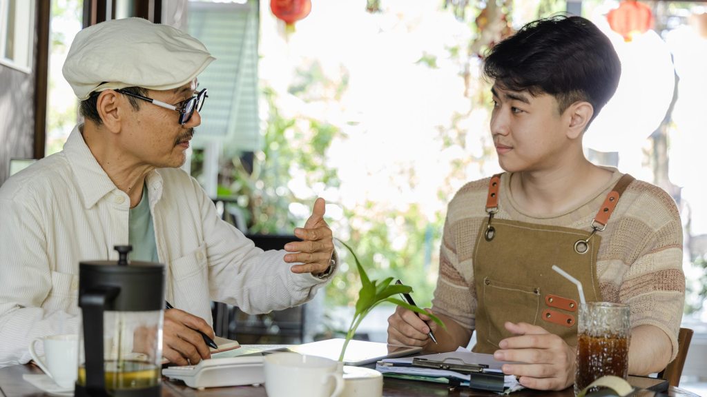An older East Asian man in a flat cap gestures while speaking to a younger man wearing an apron at a table with a clipboard, calculator, and drinks.