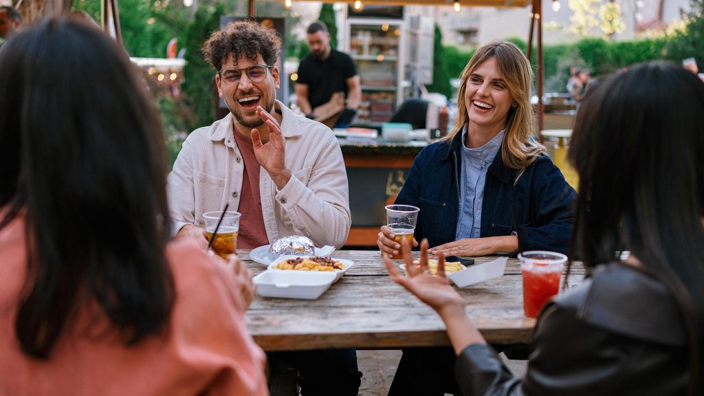 four adults sit around a rustic wooden table at an outdoor venue, engaged in lively conversation. Two people face the camera—a man with curly hair and glasses in a beige overshirt, and a woman in a dark denim jacket—both laughing and smiling broadly. The backs and shoulders of two other individuals frame the foreground. Scattered across the table are styrofoam takeout containers with loaded fries, a foil-wrapped food item, and clear plastic cups filled with beer, iced tea, and a red beverage. The softly blurred background features warm string lights and a food vendor station.